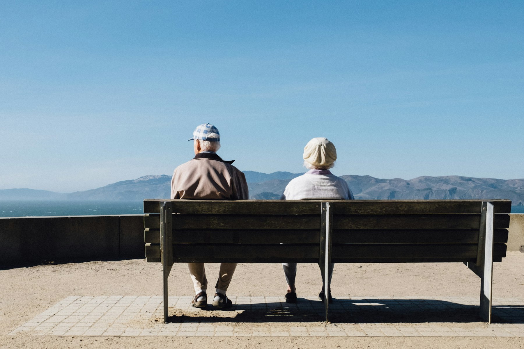 An older couple sitting together on a bench, looking out at the horizon